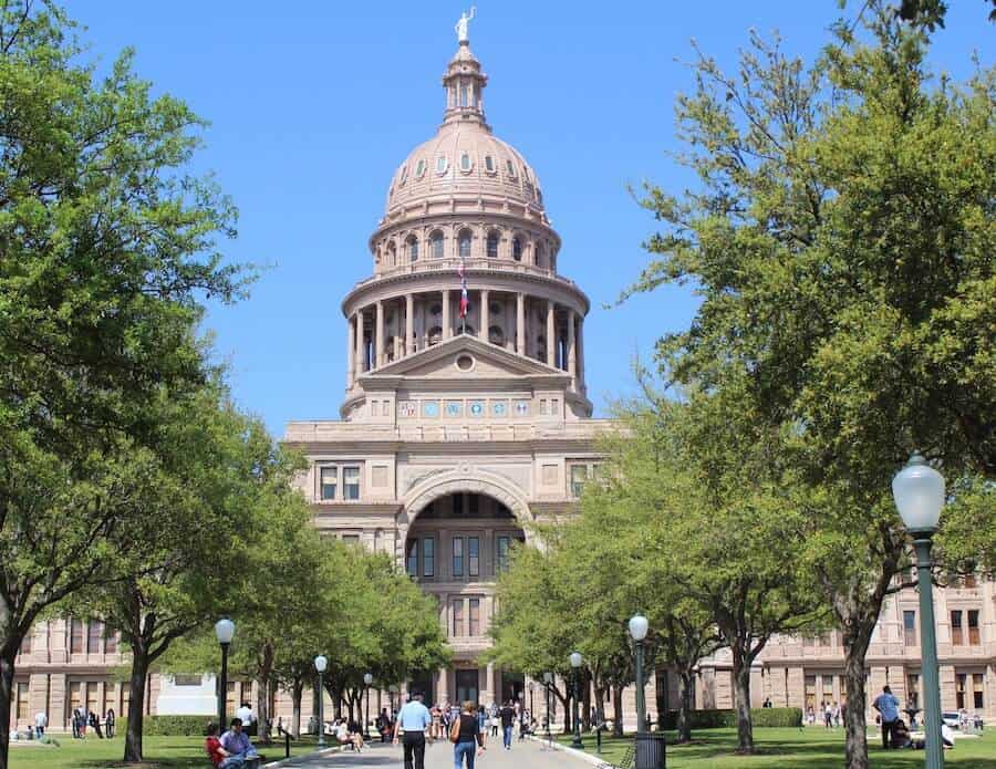 The Texas State Capitol building in Austin, Texas, stands grandly under a clear blue sky, its dome a focal point amidst the lush greenery of the surrounding trees.