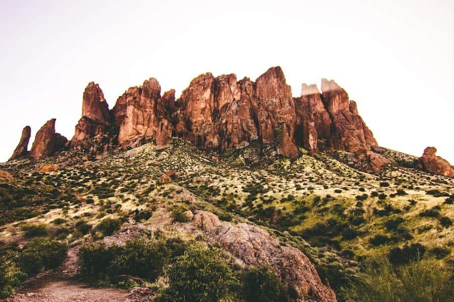 Dramatic red rock formations rising starkly from the desert landscape, dotted with green shrubbery, under a hazy sky near Phoenix, Arizona, a rugged and beautiful backdrop for college students seeking an adventurous spring break.