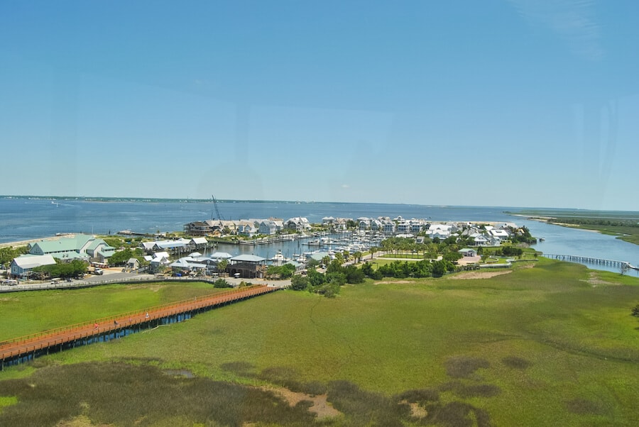 Panoramic view from Old Baldy Lighthouse overlooking the serene Bald Head Island, with its coastal homes and marina set against the backdrop of the expansive Atlantic Ocean and lush marshlands, a tranquil retreat for spring breakers seeking a peaceful escape.