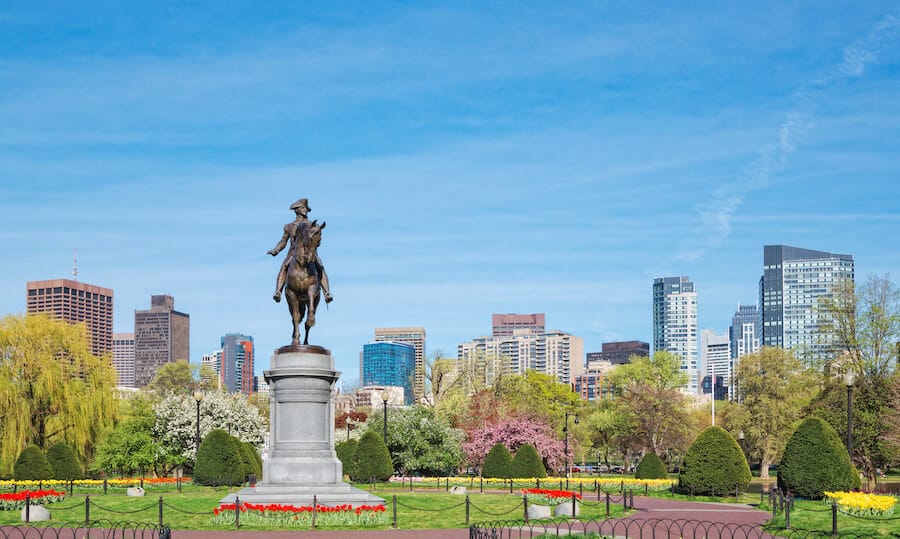 The historic Paul Revere statue takes center stage at the Boston Public Garden in Massachusetts, with lush spring blooms and manicured hedges against the Boston cityscape.