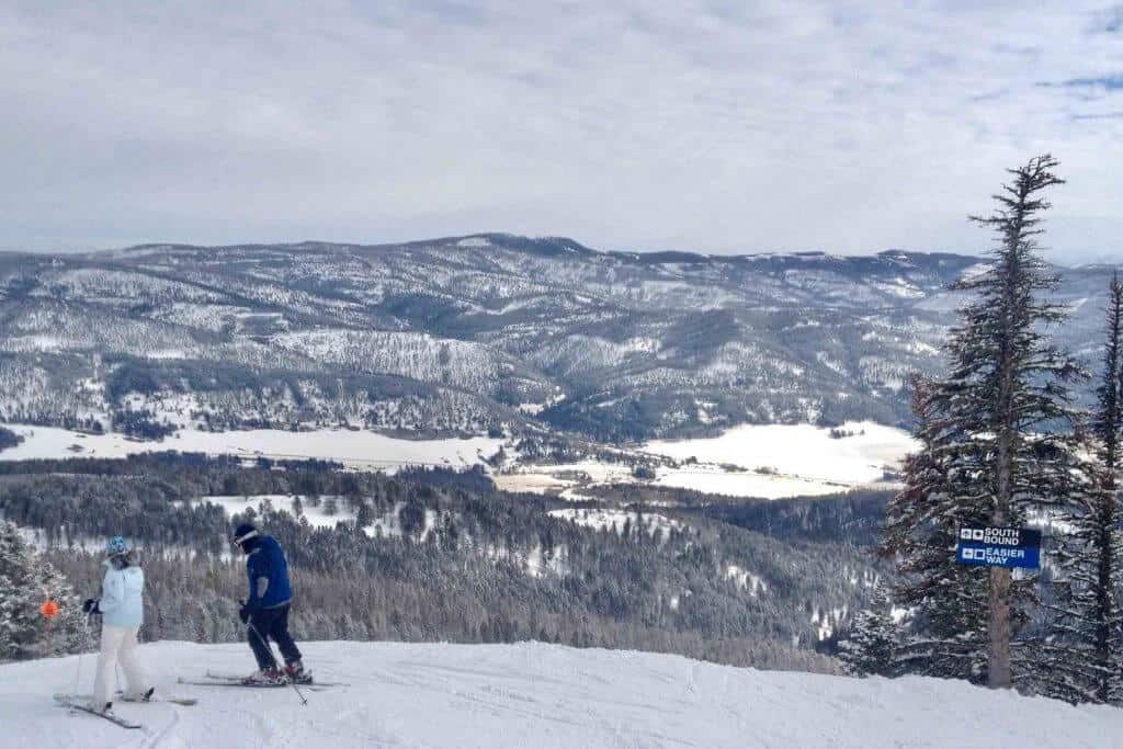 Skiers preparing to descend a snowy slope at Bridger Bowl, with panoramic views of forested mountains under a cloudy sky near Bozeman, Montana, offering a serene and thrilling spring break experience for college students.