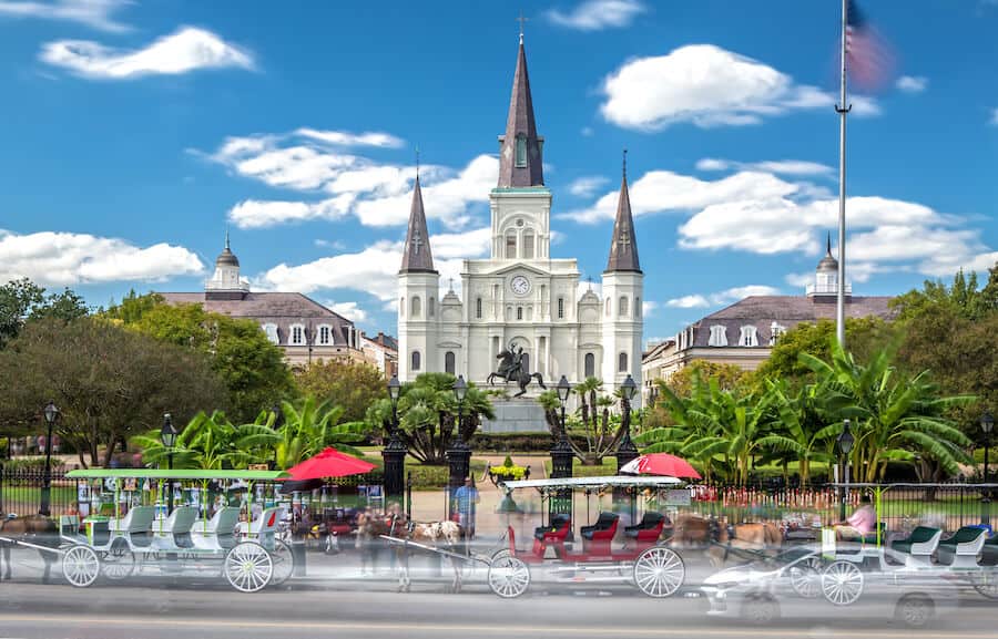 The iconic St. Louis Cathedral stands majestically against a backdrop of partly cloudy skies in Jackson Square, New Orleans, flanked by horse-drawn carriages awaiting riders, a vibrant destination for college spring breakers seeking culture and history.