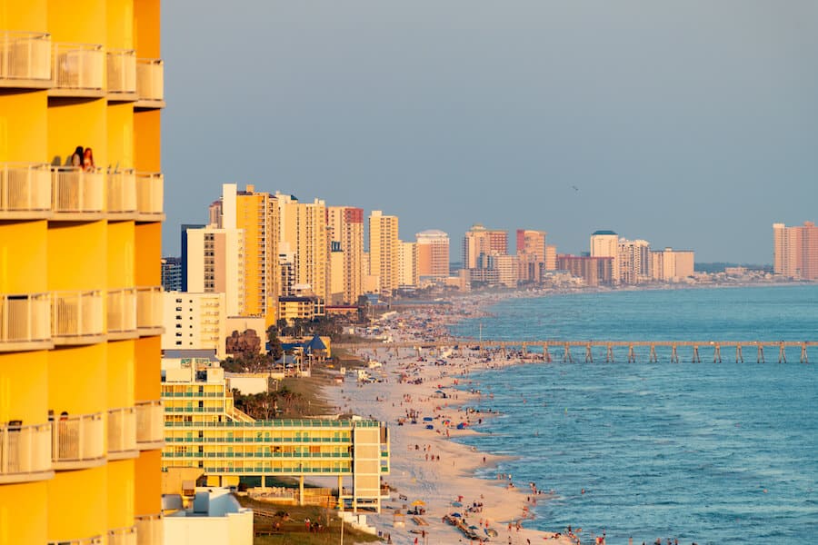 he vibrant Panama City Beach skyline, with its row of towering beachfront high-rises glowing in the golden hour sunlight, overlooks a bustling beach scene teeming with vacationers. PCB is one of the most popular destinations in the USA for college spring breakers.