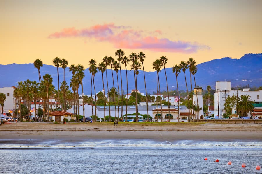Twilight descends on Santa Barbara, California, as seen from the pier with tall swaying palm trees lining the shore, the Santa Ynez Mountains in the distance, and a peaceful beachfront.