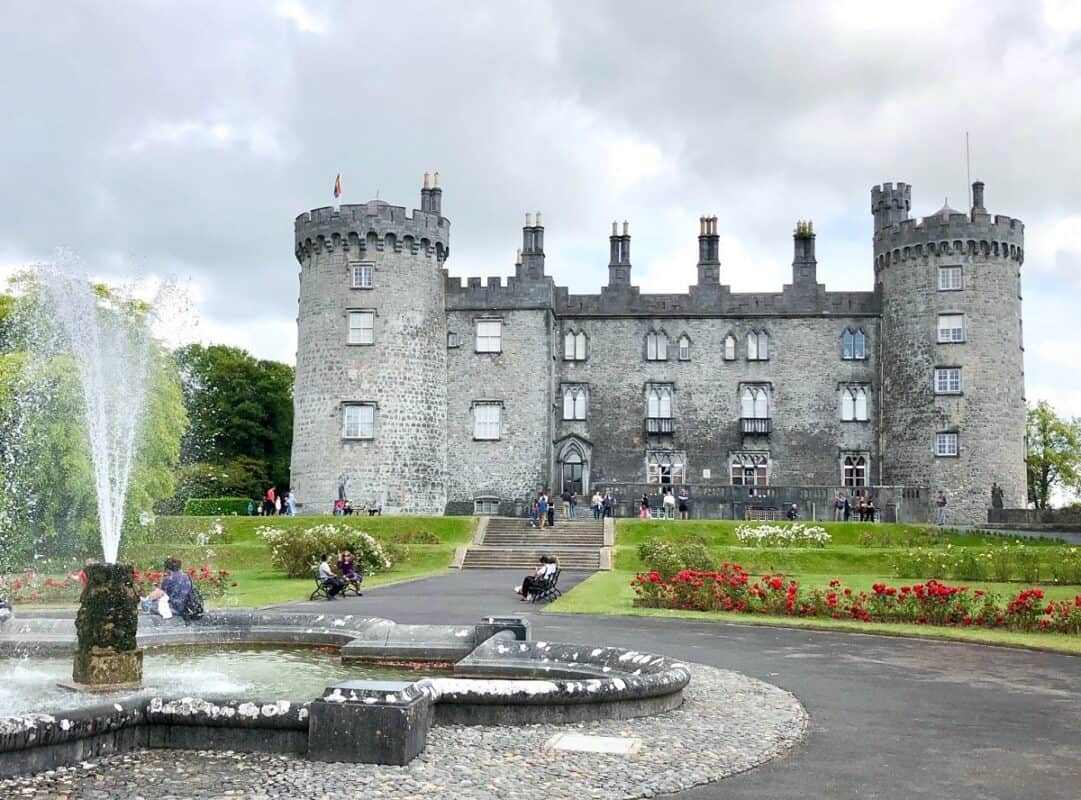 Kilkenny Castle and a fountain that sits in front of the castle, with tourists walking on the path and sitting on benches lining the path leading from the fountain to the castle