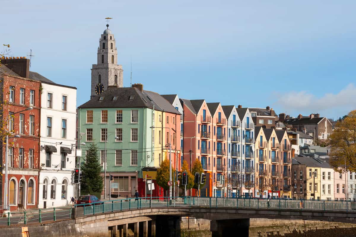 Colorful facades of row houses along a street in Cork, Ireland, with the prominent Shandon Bells tower in the background, reflecting the city's vibrant architecture and inviting atmosphere for travelers visiting Ireland on a budget.